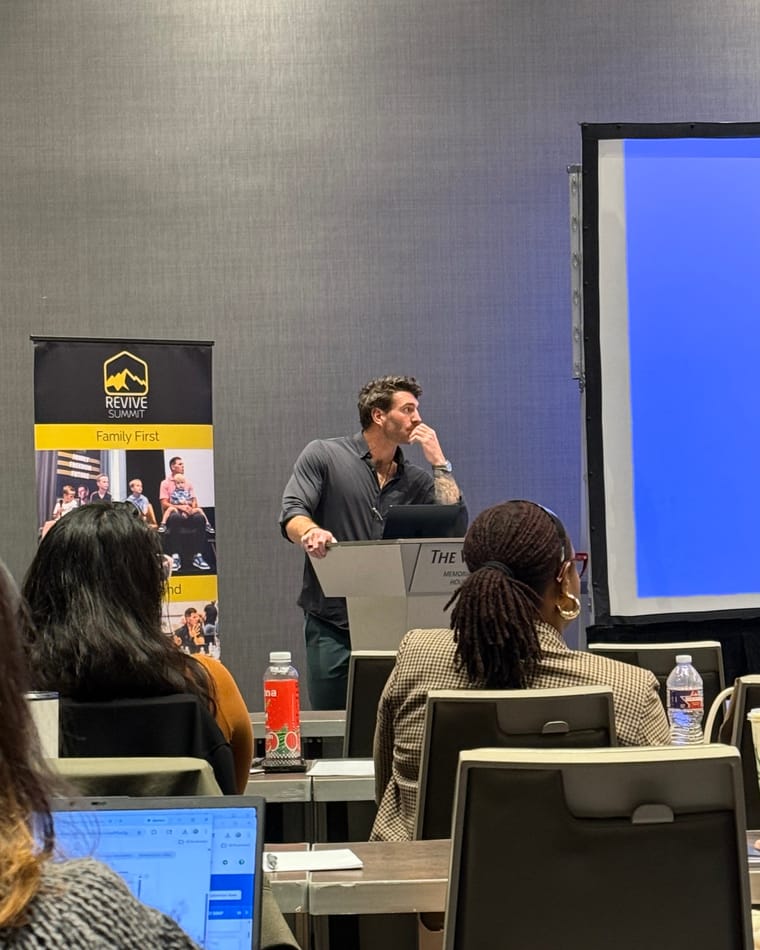 Maxwell Mayes presenting from a lectern to a seated audience at the Revive Summit.