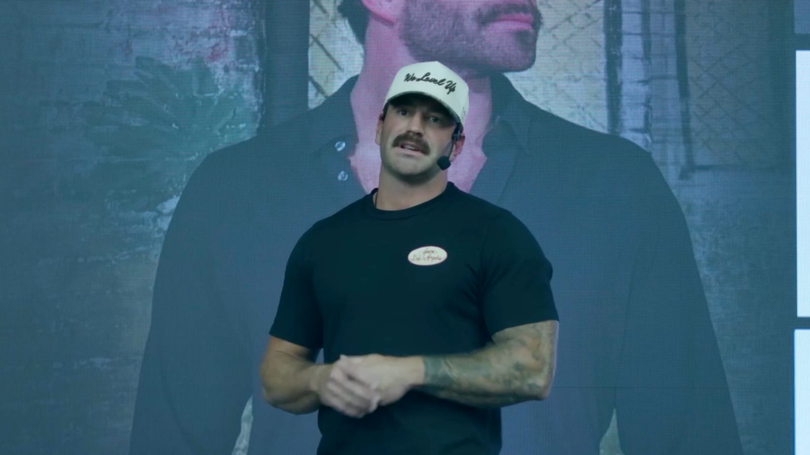 Maxwell Mayes speaking on stage in a headset microphone, in front of a large screen showing his photo.